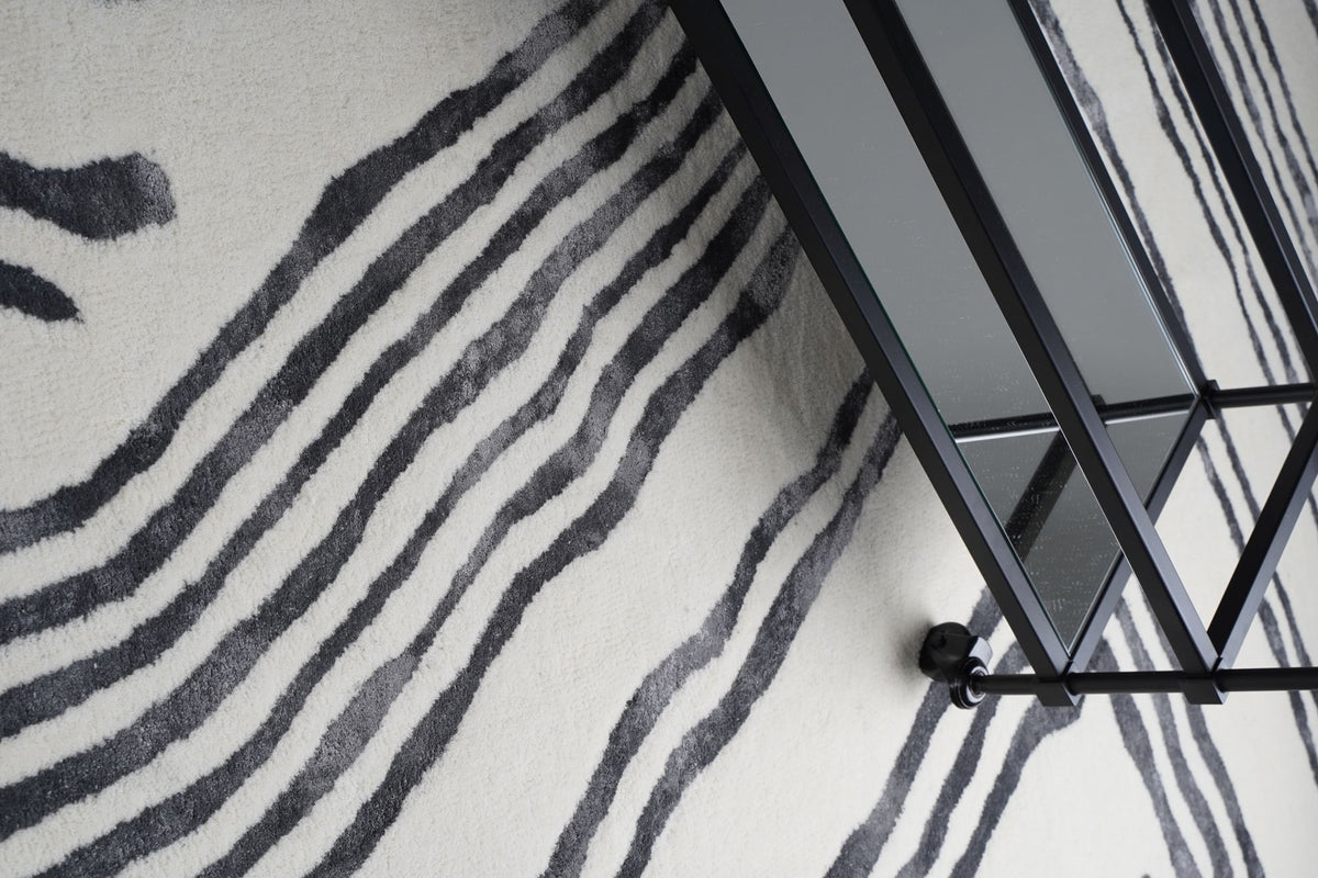 Livid Wool & Silk Rug seen from above under a black metal and glass side table, hand tufted New Zealand wool and Botanical Silk in dark flowing lines.